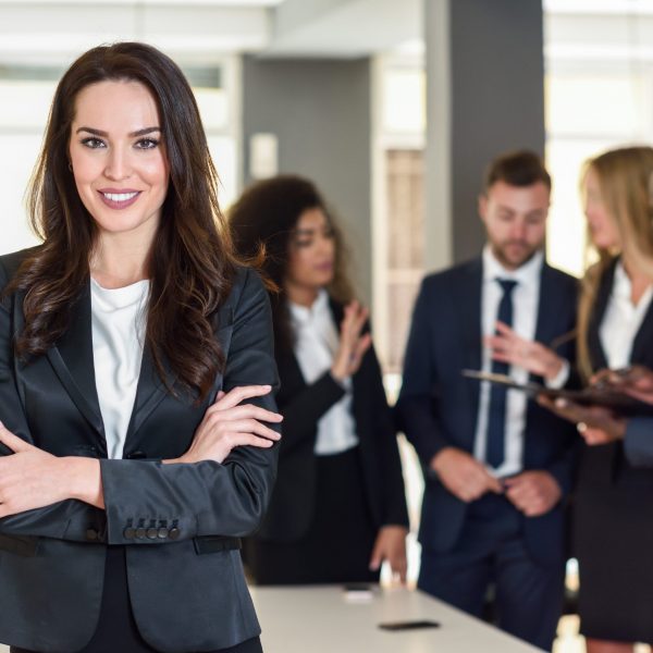 Businesswoman leader looking at camera in modern office with multi-ethnic businesspeople working at the background. Teamwork concept. Caucasian woman.